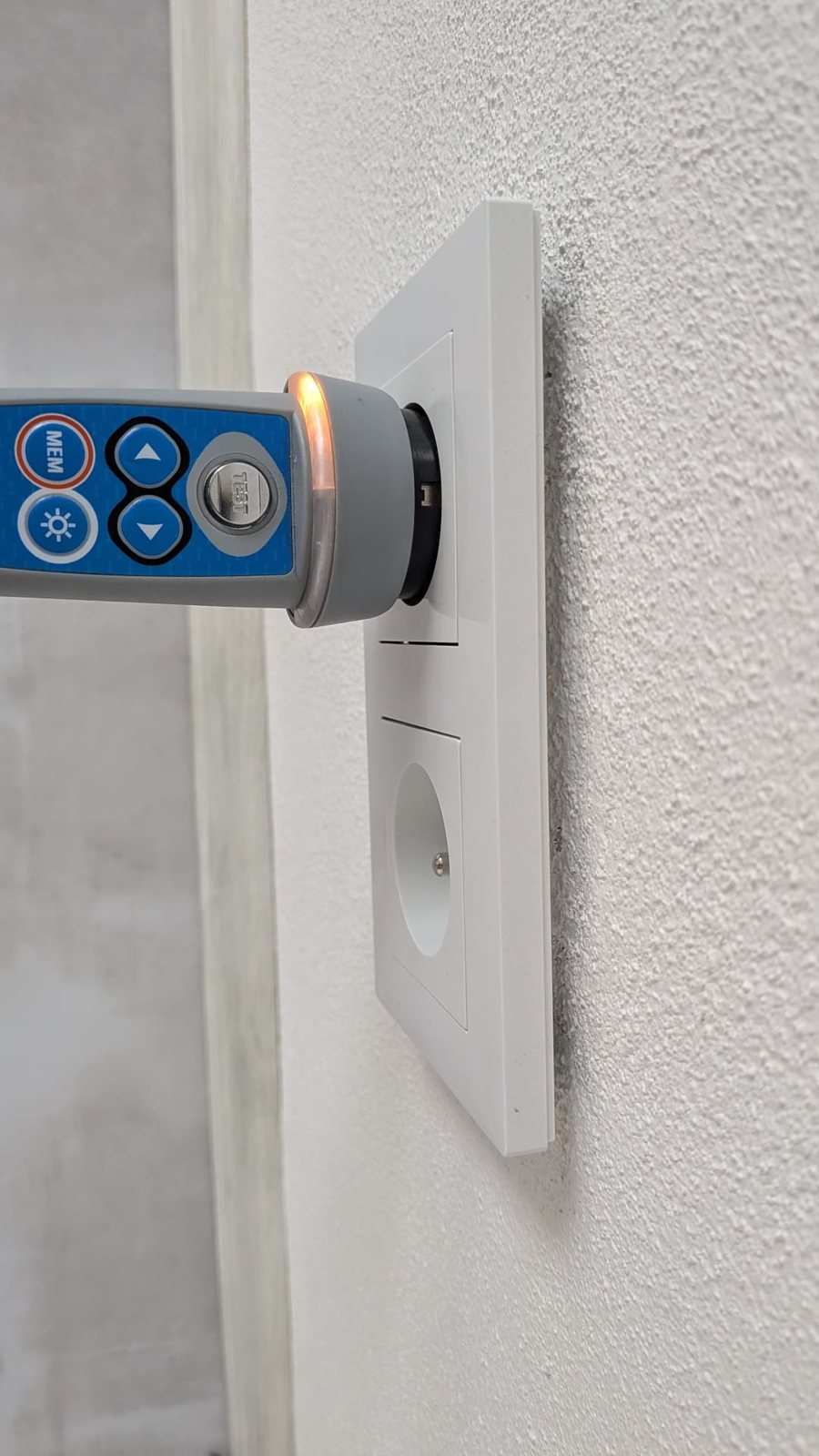 Revizní tester s kontrolkou zapojený v zásuvce na bílé stěně